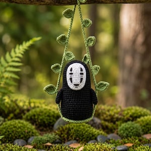 May include: A crocheted amigurumi figure of a black-bodied character with a white mask featuring black facial markings. The figure is suspended by a green crocheted vine with small leaves, hanging from a horizontal wooden branch.