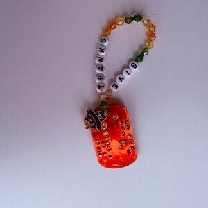 May include: An orange dog tag-shaped charm with the words "THANKS GIVE" spelled out in white letter beads. The charm has a witch hat design and is adorned with colourful beads.