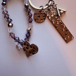 Op de afbeelding: Een zilveren sleutelhanger met meerdere bedels. De bedels zijn onder meer een ronde bedel met kleine gaatjes, een rechthoekige bedel met roze strass-steentjes en een honkbalknuppel- en balbedel. Een kralenarmband met paarse en transparante kralen is bevestigd.