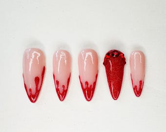 May include: A set of five almond-shaped artificial nails. Four nails have a pale pink base with red glitter dripping from the tips. The center nail is entirely covered in red glitter and adorned with red rhinestones.