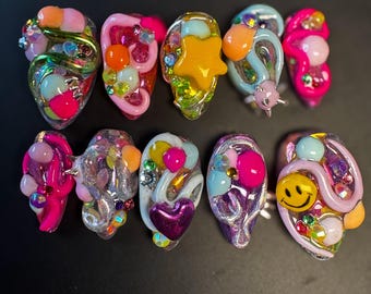 May include: A set of ten colorful, sculpted acrylic nails. Each nail features a unique design with vibrant colors, glitter, and embellishments like stars, hearts, and smiley faces. The nails have a glossy finish and are arranged on a black surface.