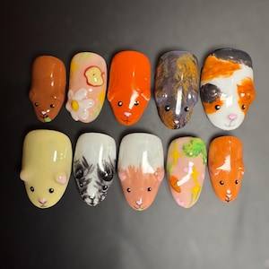 Peut inclure: Dix faux ongles décorés sur le thème du cochon d'Inde. Chaque ongle présente un design unique avec des couleurs variées, notamment marron, orange, blanc et jaune, avec des traits de visage détaillés et des motifs floraux. Présentés sur fond noir.