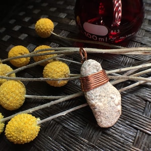 May include: A white stone pendant wrapped with copper wire. The pendant is hanging from a copper wire loop. The pendant is on a brown woven surface with dried yellow flowers.