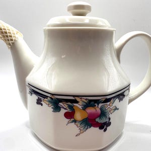 Peut inclure: Théière en céramique crème avec couvercle et anse. La théière présente une bande noire avec un motif floral et fruité jaune, rouge et violet. Le bec verseur est protégé par un filet.