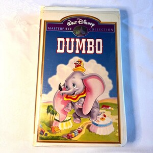 May include: A vintage VHS tape of Disney's "Dumbo" from the Masterpiece Collection. The cover features a cartoon illustration of Dumbo the elephant with large pink ears, a yellow collar, and a small circus scene in the background.
