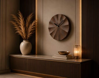 May include: A circular dark brown wooden wall clock with a sunburst-style pleated texture, featuring thin black hands. The clock is mounted on a smooth beige wall next to a cream-colored vase filled with tall, feathery dried pampas grass stems.