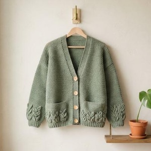 Puede incluir: Un cárdigan de manga larga en color verde salvia con cuello en V, cuatro botones de madera clara en la parte delantera y dos bolsillos. Las mangas y el dobladillo inferior presentan un patrón de punto trenzado con relieve de hojas.