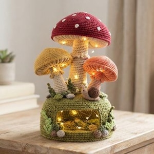 May include: A crocheted mushroom lamp featuring a large red cap with white spots, a yellow mushroom, and an orange mushroom atop a green base. The base includes a small snail, decorative foliage, and a hollow centre illuminated by warm string lights.