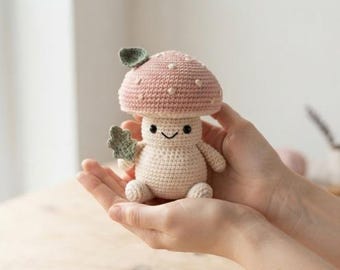 May include: A small crocheted mushroom figurine with a dusty pink cap featuring white embroidered spots. The mushroom has a cream-colored body, a simple stitched smiling face, and a small green leaf attached to its side and top.