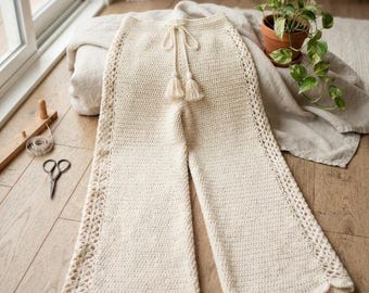 May include: Cream-colored crochet pants featuring a wide-leg silhouette, a drawstring waist with two tassels, and intricate openwork lace detailing along the side seams. The hem of the pants is finished with a scalloped edge design.