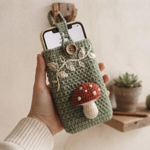 May include: A sage green crochet phone pouch featuring a three-dimensional red and white mushroom applique, embroidered white vine detailing, and a single wooden button closure. The pouch is designed to hold a smartphone securely in place.