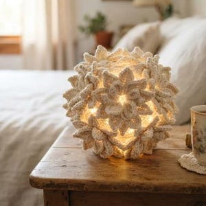 May include: A spherical lamp covered in cream-colored crocheted flower motifs that emit a warm, soft glow from within. The textured, handmade light fixture features intricate petal layers and sits on a wooden surface next to a ceramic mug.