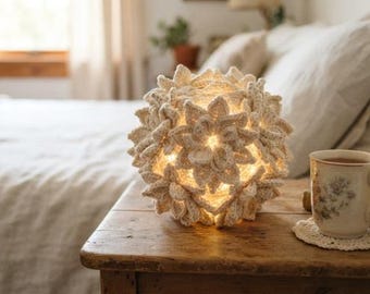 May include: A spherical lamp covered in cream-colored crocheted flower motifs that emit a warm, soft glow from within. The textured, handmade light fixture features intricate petal layers and sits on a wooden surface next to a ceramic mug.