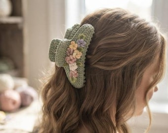 May include: A sage green hair claw clip featuring a textured crochet surface adorned with small, delicate embroidered flowers in shades of yellow, pink, and purple. The accessory is designed with a sturdy spring mechanism to hold hair securely in place.