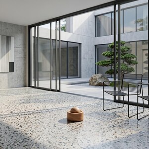 Puede incluir: Un interior moderno con grandes puertas y ventanas de cristal. La habitación presenta suelo de terrazo, paredes de hormigón y sillas de metal negro. Un cuenco de madera con una esfera está en el suelo, y un bonsái es visible afuera.