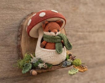 May include: A crocheted fox figurine wearing a green scarf, nestled inside a cream-colored mushroom house with a red cap featuring white spots. The structure is mounted on a circular wooden slice base decorated with green moss and small acorns.