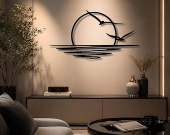 May include: A black metal wall art piece featuring a circular sun silhouette with two birds flying across it, positioned above a series of horizontal lines that create a rippled water reflection effect against a plain wall surface.