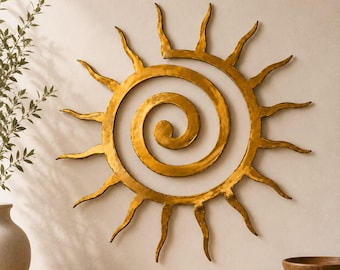 May include: A gold-colored metal wall art piece shaped like a sun with a central spiral design. The sun features numerous radiating, wavy rays extending outward from the circular core, creating a textured, metallic finish on the surface.