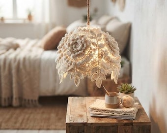 May include: A cream-colored crocheted pendant lamp shade featuring intricate floral and leaf motifs. The shade is suspended by a twisted cord and emits a warm, soft glow through its textured, open-work fabric construction.