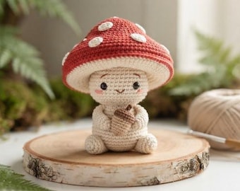 May include: A small, handcrafted crochet mushroom figure with a red cap featuring white spots, a cream-colored body, and a smiling face with pink cheeks. The figure is holding a small, light brown crocheted acorn in its hands while sitting upright.