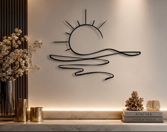 May include: A black metal wall art piece featuring a minimalist line drawing of a sun rising over a horizon with wavy lines representing water. The design consists of a semi-circle sun with radiating rays above a series of flowing horizontal lines.