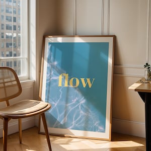 Puede incluir: Un cuadro enmarcado con la palabra "flow" en letras doradas sobre un fondo azul con efecto de ondas de agua. El cuadro está apoyado contra una pared, junto a una silla de madera y una mesa pequeña.