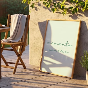 May include: A rectangular framed print with a light-coloured background and the green cursive text memento vivere. The frame is made of light-toned wood and is positioned upright on a tiled surface next to a wooden chair and a potted plant.