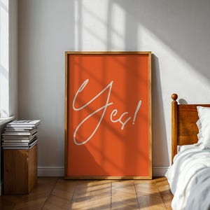 Op de afbeelding: Een oranje kunstprint met het woord "Yes!" in witte cursieve letters, ingelijst in lichtbruin. De print wordt in een slaapkamer getoond. De kamer heeft een houten vloer en een witte muur.