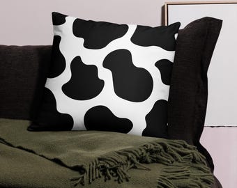 Può includere: Un cuscino quadrato con un motivo a stampa di mucca. Il cuscino presenta grandi macchie nere irregolari su uno sfondo bianco. Il cuscino è posto su un divano grigio scuro con una coperta verde.