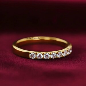 May include: A gold band ring with a row of small, clear gemstones. The ring is set against a deep red background. The ring is a simple design, with the gemstones set close together. The ring is likely made of gold.