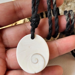 Op de afbeelding: Een ronde witte schelphanger met een natuurlijk spiraalpatroon, hangend aan een dik zwart gedraaid koord. De hanger heeft een glad, gepolijst oppervlak met subtiele crèmekleurige wervelingen en is vastgezet met een eenvoudige knoop.
