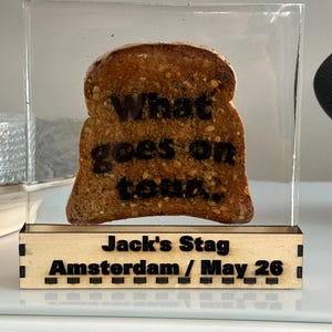Puede incluir: Un objeto decorativo con una rebanada de pan tostado en acrílico transparente. La tostada tiene la inscripción "What goes on tour". La base de madera tiene grabado "Jack's Stag Amsterdam / May 26."