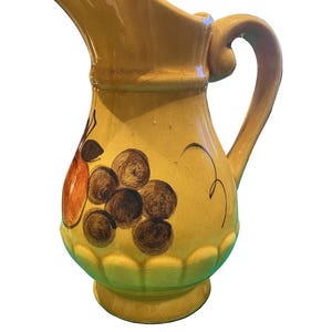 May include: A yellow ceramic pitcher with a curved handle and spout. The pitcher is decorated with a painted design of grapes and a pear. The base of the pitcher has a scalloped edge.
