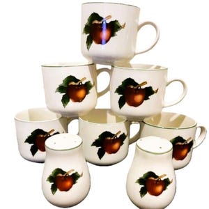 May include: A collection of white ceramic mugs and salt shakers. Each piece features a red apple design with green leaves and a green rim. The mugs have a handle, and the salt shakers have a perforated top.