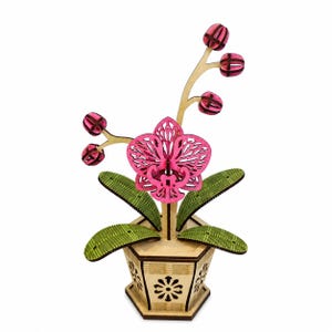 May include: A laser-cut wooden orchid sculpture featuring a bright pink flower with intricate cutouts, five small pink buds on stems, and four green leaves. The plant is set in a light-coloured hexagonal wooden pot with floral cutouts.