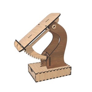 May include: A wooden phone stand with a gear mechanism. The stand is made of light brown wood with dark brown accents. It features a tray for holding a phone and a base with a small drawer.