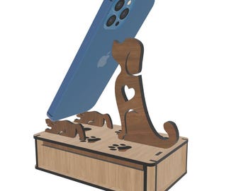 May include: Wooden phone stand with a dog design. The stand features a light brown box base, a dark brown dog silhouette with a heart cutout, and two small dog figures. A blue smartphone rests on the stand.
