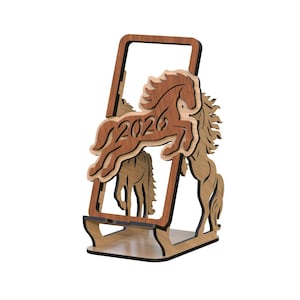 Pode incluir: Um suporte de telefone de madeira com um design de cavalo a saltar e o ano "2026" recortado. O suporte é feito de madeira em camadas em tons de castanho e bege, com uma moldura e uma base retangulares.