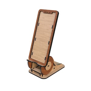 Puede incluir: Soporte de teléfono de madera con un marco en forma de teléfono marrón claro y un borde marrón más oscuro. El soporte tiene un mecanismo similar a un engranaje para ajustar el ángulo y una base rectangular. El soporte es de madera.