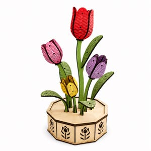 Può includere: Una scultura decorativa composta da quattro tulipani in legno a strati di colore rosso, rosa, giallo e viola, con steli e foglie verdi. I fiori sono montati su una base ottagonale in legno chiaro decorata con sagome di tulipani neri.