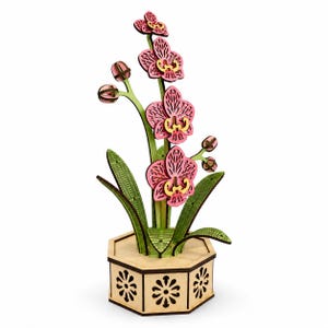 Può includere: Una pianta di orchidea decorativa in legno con petali rosa a strati con centri gialli, steli verdi e foglie verdi testurizzate. La pianta è inserita in un vaso esagonale in legno chiaro con intagli floreali marrone scuro su ogni lato.
