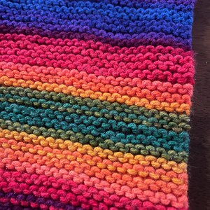May include: Close-up of a hand-knitted textile with a vibrant rainbow color scheme. The knitted fabric displays horizontal stripes of various colors, including red, orange, yellow, green, blue, and purple. The texture of the yarn is visible.