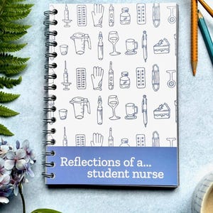 May include: A spiral-bound notebook with a white cover featuring blue line art of medical supplies. The notebook has a blue band with the text "Reflections of a... student nurse". The notebook is surrounded by art supplies and flowers.