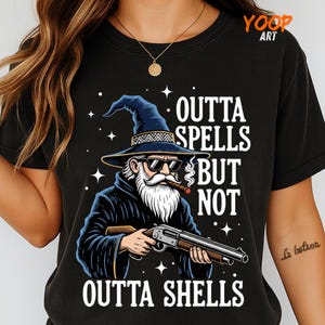 Puede incluir: Camiseta negra con un diseño de un mago con barba, gafas de sol y un cigarro, sosteniendo una escopeta. El texto dice "Outta Spells But Not Outta Shells."