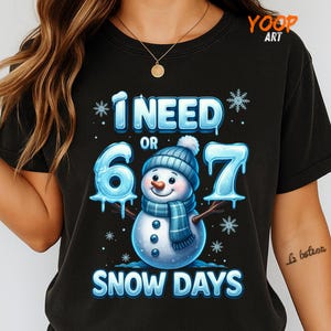 May include: Black t-shirt with a snowman graphic. The snowman wears a blue hat and scarf, and the text reads "I NEED 6 OR 7 SNOW DAYS". Snowflakes and the words are in a light blue color.