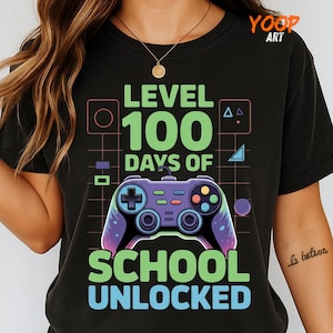 May include: Black t-shirt with a graphic design featuring a video game controller and the text "LEVEL 100 DAYS OF SCHOOL UNLOCKED" in green and blue. The design includes geometric shapes and a grid pattern.