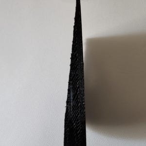 May include: A black, wedge-shaped object with a textured, layered surface. The item features a sharp, pointed tip at the top and a wider, flat base at the bottom, displaying a distinct 3D printed finish with visible horizontal lines throughout.