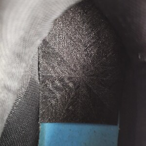 May include: The interior of a shoe featuring a black heel insert with a textured, cross-hatched pattern. Below the black insert, a bright blue foam insole is visible. The surrounding lining of the shoe is a dark gray, textured fabric material.