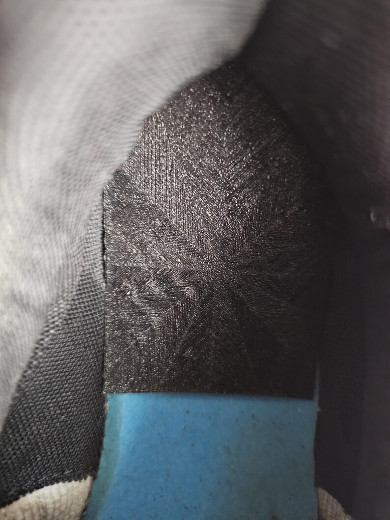 May include: The interior of a shoe featuring a black heel insert with a textured, cross-hatched pattern. Below the black insert, a bright blue foam insole is visible. The surrounding lining of the shoe is a dark gray, textured fabric material.
