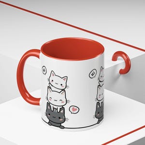 May include: A white ceramic mug with a red interior and handle. The mug features a stack of cartoon cats in white and gray, with a heart and the word "Hi" in speech bubbles. The mug is a fun and whimsical item.